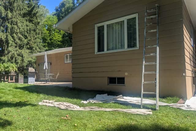 A tan ranch house with a ladder against the wall and drop cloths spread on the grass.