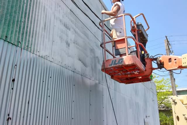 A painter spraying a large metal warehouse wall from a boom lift basket.